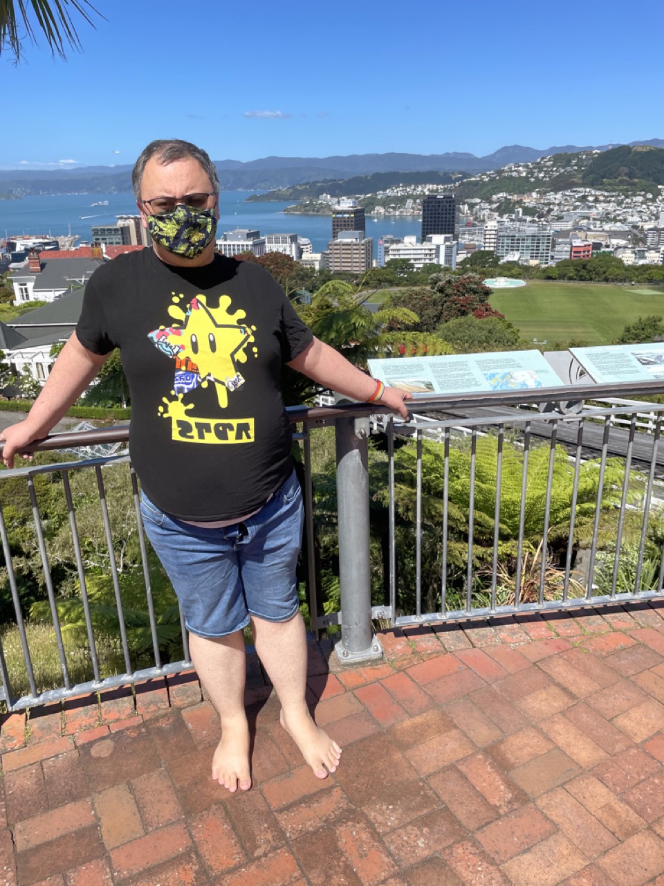 Feets leans against a fence railing outside of the cable car. Wellington city on a clear, sunny day can be seen behind in the distance