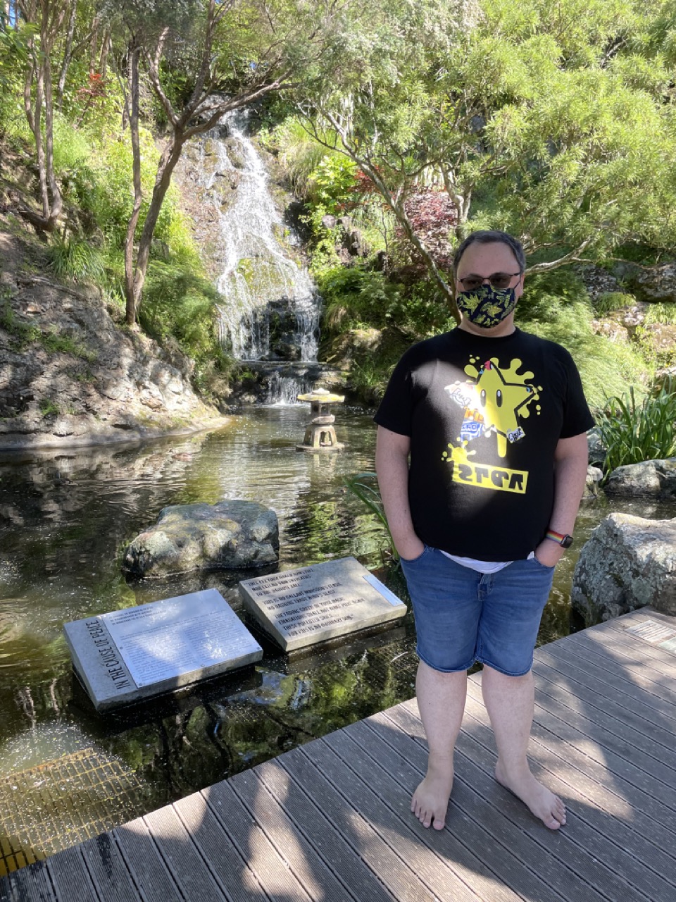Feets stands in the shade near a pond in the middle of which is a small, Japanese-style lantern which has no flame. A waterfall and some green trees surround the frame of the picture