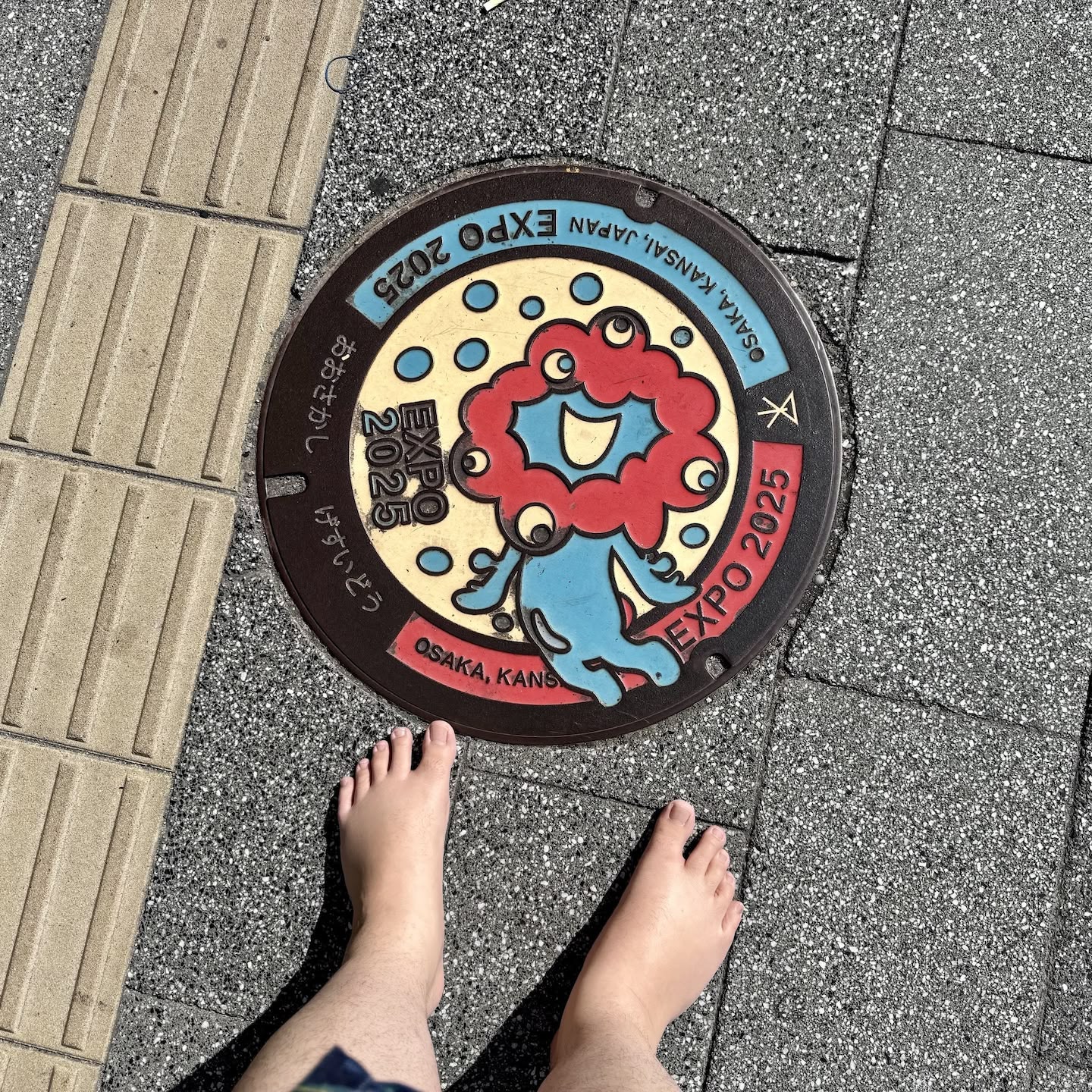 A picture of an Ōsaka Expo 2025-themed manhole cover with the multi-eyed blue and red mascot on it. The tops of my bare feet are visible in the shot as I took the photo pointing directly down.