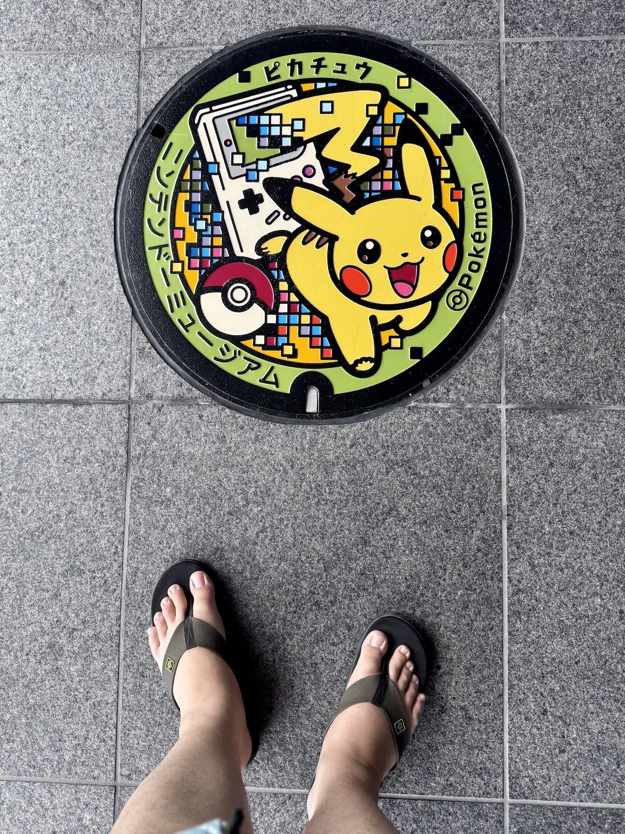 A picture of a Pokémon-themed manhole cover with Pikachu running from a Game Boy and a Pokéball. The tops of my feet, wearing jandals, are visible in the shot as I took the photo pointing directly down.
