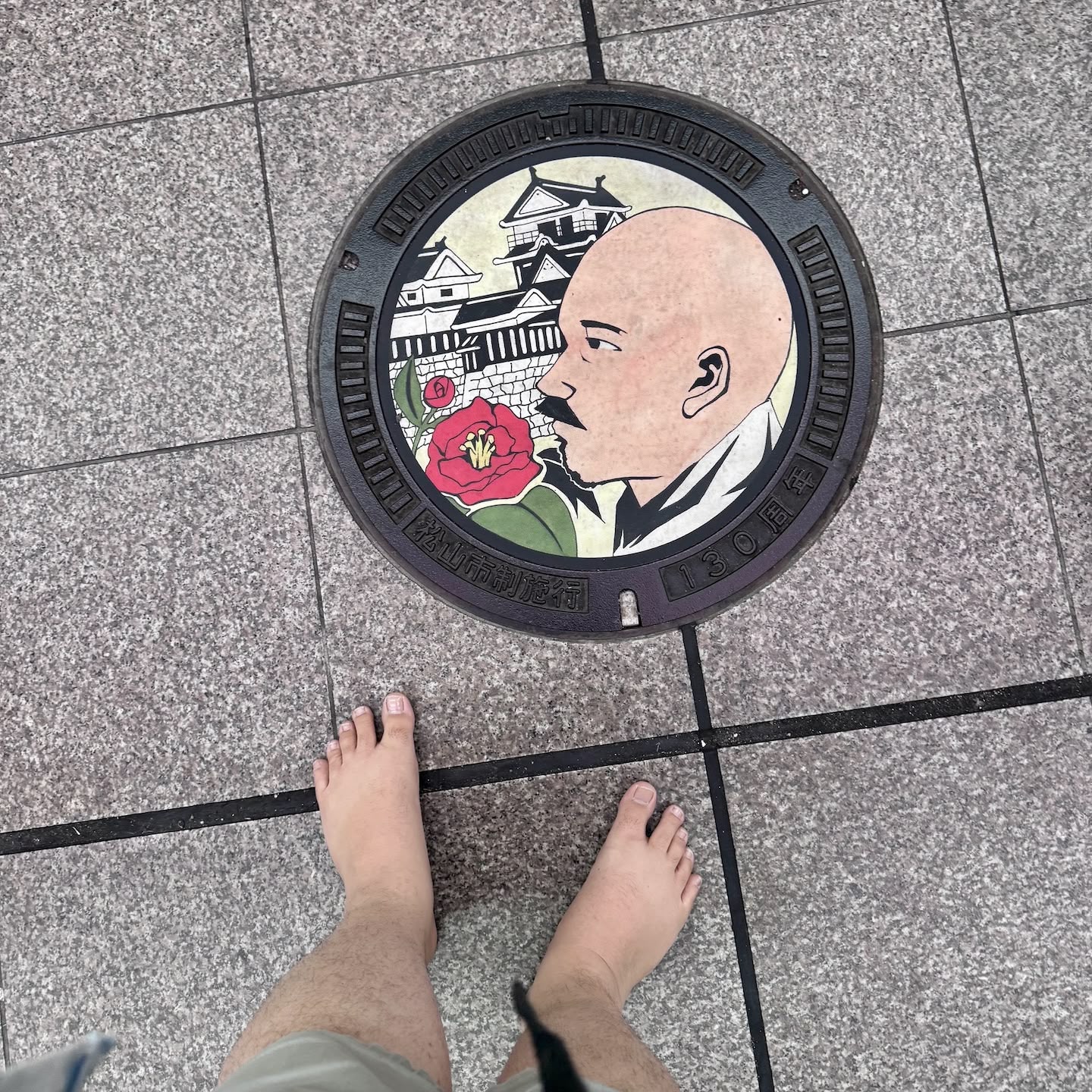 A picture of a Matsuyama-themed manhole cover with a depiction of Masaoka Shiki, a local poet from years ago, next to a Camellia flower in front of Matsuyama Castle. The tops of my bare feet are visible in the shot as I took the photo pointing directly down.