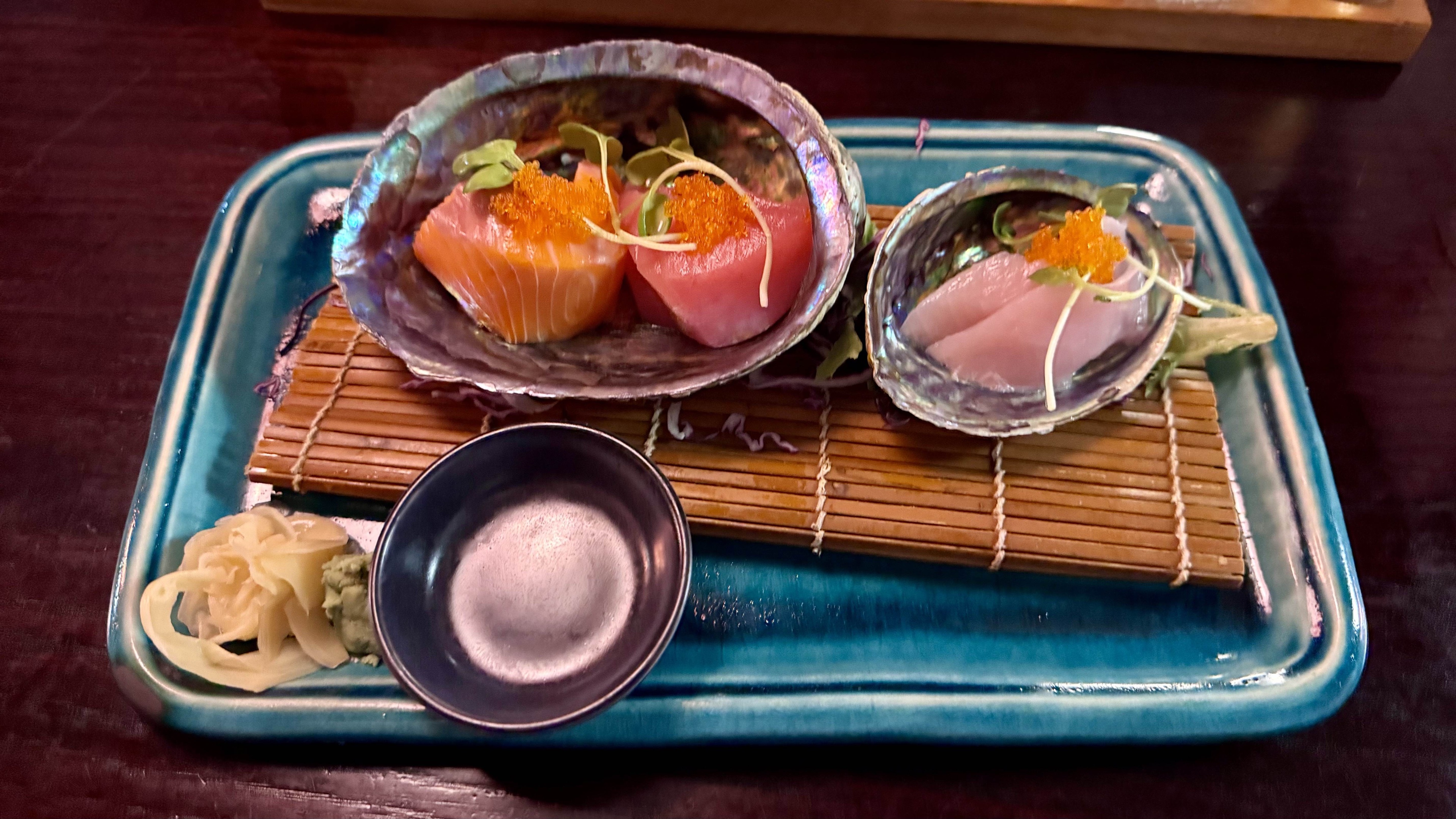 Salmon, tuna, and white fish sashimi served on pāua shells, themselves on a blue platter with ginger and wasabi.